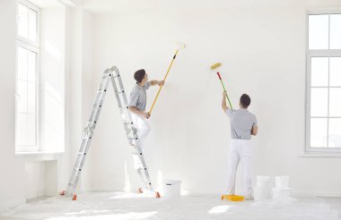 Üniformalı iki erkek ressam bir duvarı silindirle beyaza boyar. Ev onarımı konsepti. Müteahhitlerin profesyonel işi. Modern apartman dekorasyonu, oturma odası iç dekorasyonu.