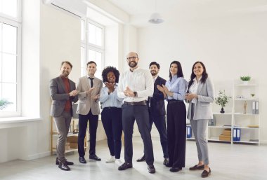 Farklı etnik çeşitlilikte iş yapan bir grup erkek ve kadın kameraya bakıp ofiste alkışlıyor ve gülümsüyor. Şirket çalışanları ya da bir grup personel iyi bir anlaşma yapmaktan mutluluk duyar..