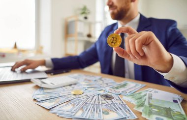 İş adamı, takım elbiseli, ofiste dizüstü bilgisayarla çalışıyor, elinde bir Bitcoin tutuyor, online mali işlemleri ve ticareti sembolize ediyor, şifreli para ve dijital parayı sembolize ediyor..
