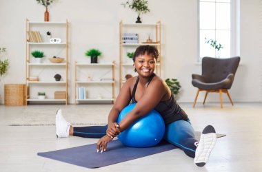 Mutlu, genç, gülümseyen, Afro-Amerikan kadın spor giyimli evde spor egzersizleri yaptıktan sonra formda bir topla yoga minderinde oturuyor. Spor, spor ve vücut pozitif konsept.