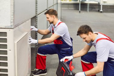 Üniforma giyen profesyonel elektrikçiler, çatıdaki ağır vasıflı merkezi havalandırma sistemini aletlerini kullanarak tamir veya onarıyor. HVAC 'yi tamir eden erkek işçiler ya da mühendisler.