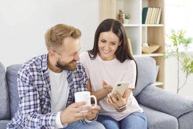 Mutlu gülümseyen genç çift cep telefonunu birlikte kullanıyor, sosyal medyada sörf yapıyor, e-bankacılık yapıyor, e-ticaret yapıyor ya da evdeki oturma odasındaki kanepede oturup başvuru üzerinde online alışveriş yapıyor..