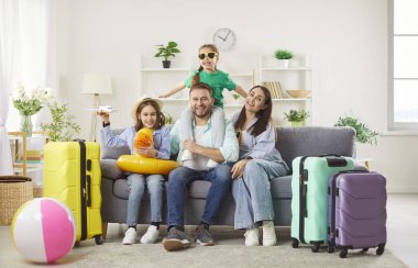 Çocukları olan mutlu ebeveynlerin portresi. Kanepede bavullarıyla oturuyor ve birlikte tropik tatil köyüne uçmaya hazır kameralara bakıyor. Aile tatili ve tatil gezisi.