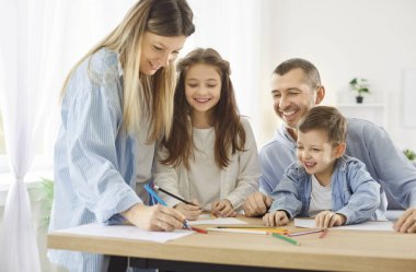 Mutlu gülümseyen çocuklar, aileleri evde oturma odasında oturmuş renkli kalemlerle resim çiziyorlar. Çocukların erkek ve kız kardeşleri albümde resim yapıyor. Aileyle vakit geçiriyorlar..