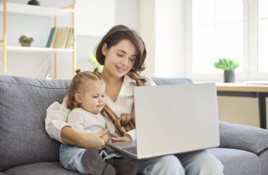 Mutlu anne ve küçük kızı evdeki oturma odasındaki kanepede dinleniyor, online eğlence için dizüstü bilgisayar kullanıyorlar. Bu aile anında internet bağlanması, boş zaman ve kaliteli dinlenme zamanı vurgulanıyor..