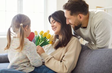 Mutlu anne evde, oturma odasındaki kanepede oturuyor. Tatil kutlamaları sırasında elinde çiçek, elinde de kız ve koca laleleri var. Aileden hediye alan mutlu kadın..