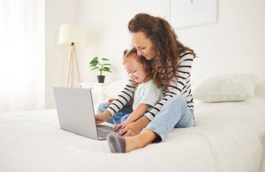 Gülümseyen genç anne, küçük kızıyla büyük beyaz bir yatakta oturuyor ve dizüstü bilgisayar kullanıyor. Mutlu aile babaları ve çocukları hafta sonları birlikte vakit geçiriyorlar, eğleniyorlar ve eğleniyorlar..