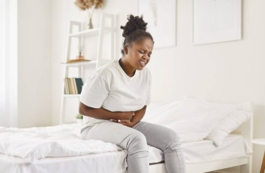 Hasta Afro-Amerikan kadın yatakta oturuyor ellerini karnına koyuyor, şiddetli karın ağrısı çekiyor, mutsuz bir kız karın ağrısı çekiyor, pankreatit gastrit semptomu, apandisit. 