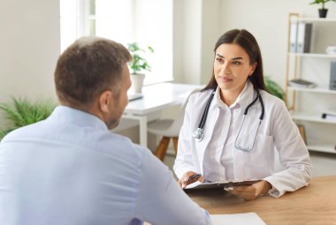 Kadın doktor ya da hemşire, hastane muayenehanesinde erkek hastaya danışır. Hasta randevusu sırasında tıbbi destek ve doktor bakımına odaklanın.