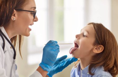 Klinik ziyareti sırasında hastanın boğazını muayene eden doktor, elinde tahta spatulayla gezen çocuk doktoru iltihaplı bademcikleri, burun solunumunu inceleyen, profesyonel tıbbi yardım sağlayan.