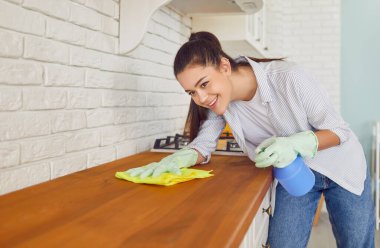 Plastik eldivenli, gülümseyen genç bir ev hanımı mutfak tezgahının yüzeyini temizliyor deterjanla temizliyor. Ev işleri ya da ev işleri ve temizlik hizmeti konsepti.