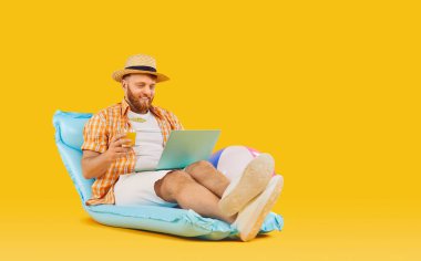 Pozitif adam tatilin tadını çıkarıyor. Portakal rengi bir arka planda dizüstü bilgisayar ve kokteyl eşliğinde şişme bir yatakta dinleniyor. Yaz yolculuğunun ve dinlenmenin özünü kucaklıyor..