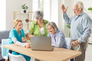 Bir pansiyondaki bir grup neşeli yaşlı insan bir dizüstü bilgisayarla çevrimiçi iletişim kuruyor. Hemşire yaşlı yetişkinlerin sosyal medyayı kullanmasına yardım eder. Dijital okuryazarlık kavramı, yaşlı yetişkinler, yaşlı ebeveynler