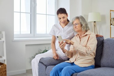 Bakıcı, internet teknolojisi olan yaşlı bir kadına telefon kullanarak yardım ediyor. Bakıcı, internet ve uygulamaların nasıl kullanılacağını göstererek, hasta desteği ve rehberlik sağlayarak destekliyor.