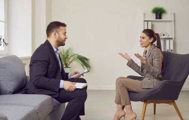 Takım elbiseli genç bir gazetecinin portresi. Elinde mikrofonla içeride sandalyede oturan çekici, gülümseyen bir kadınla röportaj yapıyor. Erkek, ofisinde kadın konuğuyla konuşuyor..