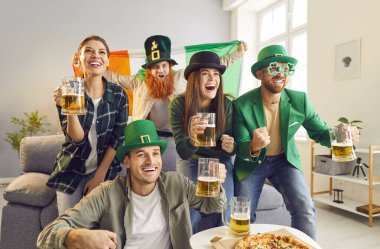 Heyecanlı yeşil şapkalı mutlu arkadaşların portresi TV 'de maç seyrediyor bira ve pizzayla evde favori takımı destekliyor ve Aziz Patrick Günü' nü kutluyor..
