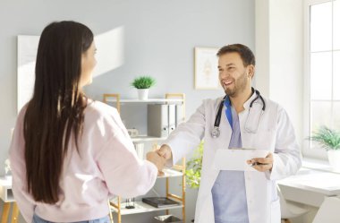 Beyaz önlüklü gülümseyen doktor, muayenehanedeki kadın hastayla el sıkışıyor. Profesyonel sağlık hizmeti sağlayıcısı, ziyaret sırasında hastayı sıcak bir şekilde karşılıyor, güven ve bağlantı kuruyor.