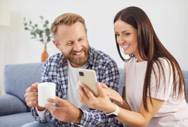 Aile çifti kanepede oturur, evde birlikte vakit geçirmenin tadını çıkarırlar. Telefon kullanarak, gülerek ve eğlenceli bir internet aktivitesiyle çevrimiçi bir eğlenceyi paylaşın.