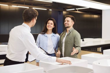 Tesisatçı dükkanındaki mutlu çift beyaz seramik lavabo seçiyor. Alışveriş merkezindeki bir aile banyo lavabosu alır. Satıcı tesisat malzemeleri satıyor. Tamir konsepti, tesisat değişimi.