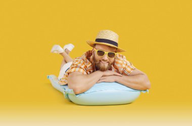 Güneş gözlüklü ve yaz kıyafetleriyle turuncu arka planda şişme bir yatakta yatan mutlu bir adamın portresi. Yaz tatilinin, dinlenmenin ve sahilde tatil eğlencesinin özünü temsil ediyor..