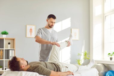 Doktor ya da fizyoterapist, kanepedeki genç adama bacak tedavisi ve fizik tedavi sağlıyor. Profesyonel terapist, erkek hastaya diz yaralanmasından sonra rehabilitasyonda yardım ediyor..