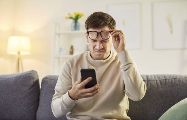 Gözlük takan genç bir adam telefonuyla miyop gözüyle mesaj okumaya çalışıyor. Prebyopia hastası bir erkek cep telefonu ekranına bakıyor. Görüş sorunları kavramı.