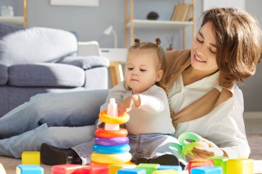 Mutlu bir anne ya da genç bir bebek bakıcısı. Küçük kızıyla piramit oyuncağı oynuyorlar. Evde sıcak bir zeminde oturuyorlar.