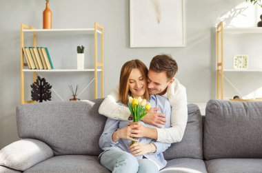 Sevgi dolu gülümseyen koca, mutlu karısını tebrik ediyor. Bir buket laleyle kanepede oturuyor ve arkasından sarılıyor. Mutlu aile çifti Uluslararası Kadınlar Günü 'nü ya da birlikte doğum gününü kutluyor..