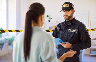 Polis memuru elinde not defteri tutarken, aydınlık odada güvenlik bandıyla kadın tanıkla konuşuyor. Soruşturma yürüten erkek polis ya da içeride polis soruşturması.
