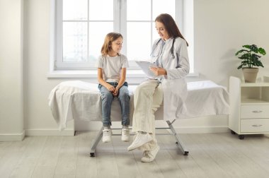 Klinikteki muayene odasında çocuk ve çocuk konuşuyor. Gülümseyen hasta çocuk ve not defteriyle genç bayan doktor yatağa oturmuş tıbbi anket dolduruyorlar.