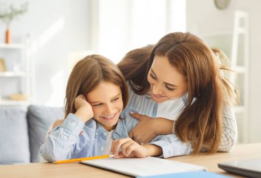 Şefkatli bir anne, gülümseyen çocuğunun üzerine eğilip destek ve cesaret veriyor. Neşeli çocuk masa başında oturup ödevlerini bitirmeye odaklanmış, sıcak ve pozitif öğrenme atmosferinin tadını çıkarıyor..