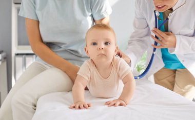 Dost canlısı gülümseyen genç bayan pediatrist, tıbbi klinikteki kanepede steteskoplu küçük şirin bebeği muayene ediyor. Anne ve çocuk muayene odasında doktoru ziyaret ediyorlar..