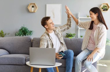 Dizüstü bilgisayar ve cep telefonuyla koltukta oturan mutlu aile çifti başarılı internet alışverişi veya online bankacılık işlemleri sonrasında birbirlerine beşlik çakıyor