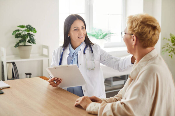 Young woman doctor in white lab coat holding clipboard, consulting with older female patient in bright medical office. Friendly cardiologist providing support and discussing health recommendations.