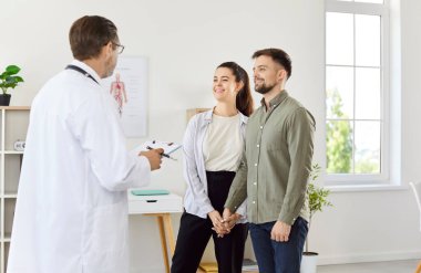 Gülümseyen genç çift koca ve karısı, klinikte ya da hastane planlamasında erkek bir aile doktoru terapistiyle konuşuyor. Sağlık hizmetleri. tıp ve kontrol konsepti.