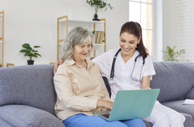 Gülümseyen genç kadın hemşire ya da doktor yaşlı bir kadına evdeki kanepede otururken dizüstü bilgisayar kullanmasında yardım ediyor. Emekliler için tıp, sağlık ve evde bakım kavramı.