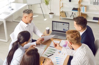 İş toplantısı sırasında veri analizi yapan iş ekibi. Ofis masasında birlikte oturup profesyonel pazarlama araçlarını tartışan, dizüstü bilgisayar kullanan ve kahve içen bir grup insan.