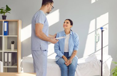 Dost canlısı doktor, vitamin tedavisi gören kadın hastalara destek oluyor. Önlüklü erkek hemşire, tıp merkezindeki yatakta oturan, damarlarında sıvı ilaç iğnesi olan mutlu bir kadınla konuşuyor.
