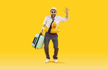 Güneş gözlüklü, lastik yüzüklü ve bavullu stüdyo sarı arka planda ofis kıyafetleri giyen genç bir adamın tam vücut fotoğrafı. Komik turist el sallayarak yaz tatiline çıkıyor..