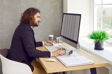 Side view of a young stylish bearded business man working on a laptop with tables and charts at the office or at home and making calculations. Business accounting, data analysis and finance concept. clipart