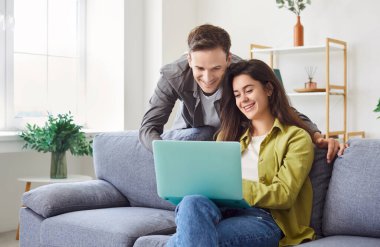 Genç bir çift evde oturuyor, kanepeye bakıyor dizüstü bilgisayara gülümsüyor, mutlu genç evli eş internette sörf yapıyor, online uçuş biletleri alıyor, iyi haberler okuyor, boş zaman geçiriyor.
