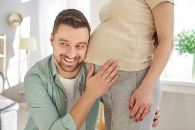 Mutlu ve yakışıklı bir adamın portresi. Güzel hamile kadınların karnını dinliyor ve evde gülümsüyor. Karısının hamileliğinin tadını çıkaran sevgi dolu bir koca. Bebek bekleyen bir çift..