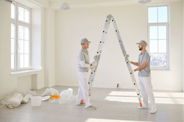 Gülümseyen neşeli boyacı takımı boş bir odada merdivenin yanında duruyor ve yeni dairenin duvarını boyamaya hazırlanıyor. Müteahhitler tadilat yapıyor. Taşınmaya hazırlanıyorlar..