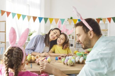 Mutlu Paskalyalar. Neşeli aile masanın etrafında toplanmış Paskalya yumurtalarını boyamak gibi eğlenceli bir aktiviteyle uğraşıyorlar. Tavşan kulaklı iki çocuk ve aileleri boya ve fırçalarla birlikte çizerler..