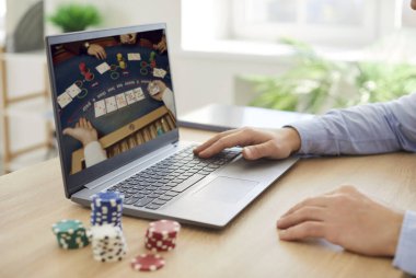 Ahşap masada oturmuş, bilgisayarında poker oynarken çekilmiş görüntüler. Elleri klavyenin üzerinde renkli poker çipleriyle çevrili sanal kumar ya da oyun deneyimi olan biri..