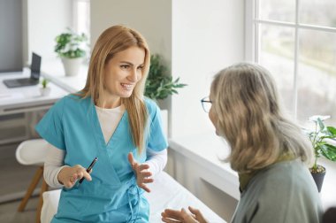 Tıp bürosundaki yaşlı, gri saçlı bir kadının portresi klinikte oturan ve muayene sırasında danışmanlık yapan neşeli kadın doktor ya da hemşireyle konuşuyor. Tıp konsepti.