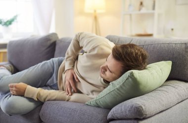 Karnına dokunan genç hasta bir adam mide ağrısından muzdarip. Evde, oturma odasındaki kanepede karın ağrısı çeken günlük giysiler giymiş bir erkek. Sağlık sorunu ve tıp konsepti.