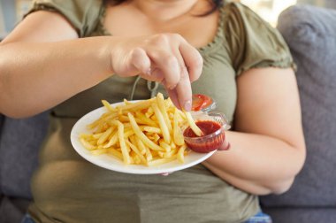 İştahla patates kızartması yiyen şişman kadın. Şişko kadının kanepeye oturup tabağı tutarak ketçapa patates kızartması bandırması. Sağlıksız yemek ve yüksek kalorili diyet konsepti