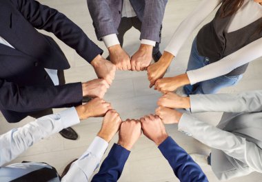 Bir grup profesyonel daire şeklinde bir araya gelerek, parlak bir ofis ortamında yapılan iş toplantısında takım çalışmasını ve birliği kutlamak için yumruk tokuşturuyor..