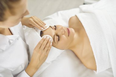 Kozmetikçi ya da güzellik uzmanı yüz bakımı için güzellik salonuna gelen mutlu genç bayanın makyajını silmek için pamuk pedleri ve temizleyici süt veya mikiler suyu kullanır. Cilt bakımı kavramı